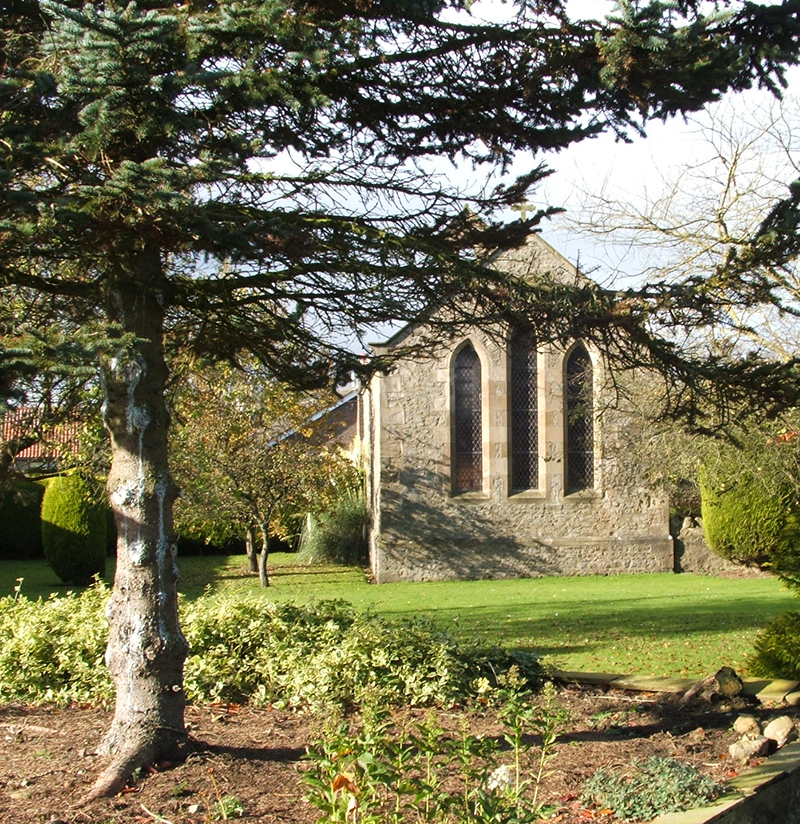 The east end of a stone-built Victorian chapel of ease with three pointed lancet windows, in a plot of land without headstones and with trees.