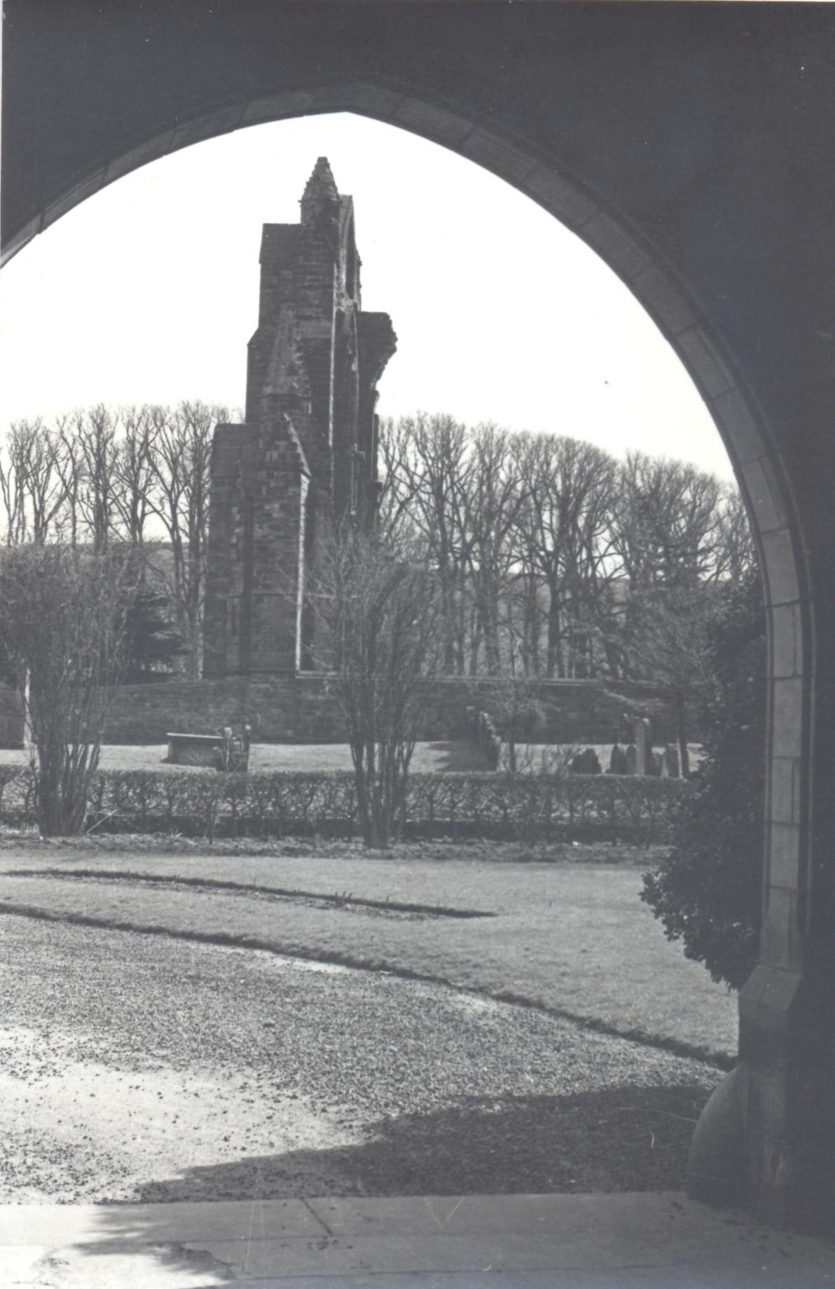 Black and white photograph. The ruins of the east end of a priory church viewed through a stone and terracotta arch with a garden and then a graveyard in between. 