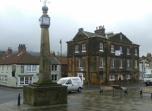 A view of Guisborough showing the Town Hall, a three storey sandstone building in a plan neoclassical style, and the market cross, an obeslisk with a sundial and weathervein on top 
